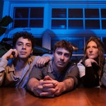 A colour photo shows four-piece group Sunglaciers sitting at a wooden table with their arms resting on top. A blue wall with windows sits behind them. 