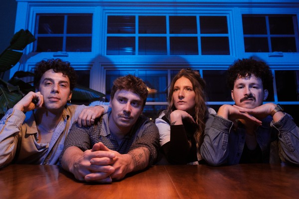 A colour photo shows four-piece group Sunglaciers sitting at a wooden table with their arms resting on top. A blue wall with windows sits behind them. 