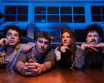 A colour photo shows four-piece group Sunglaciers sitting at a wooden table with their arms resting on top. A blue wall with windows sits behind them. 