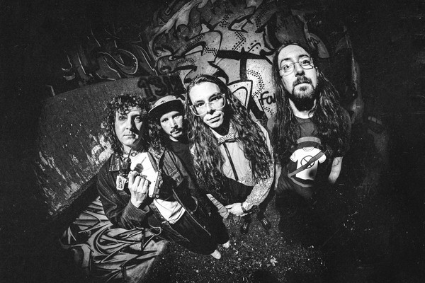 A black and white photo shows the group Tall Mary standing in front of a graffitti wall, each staring into the camera which has a warped vignette effect.