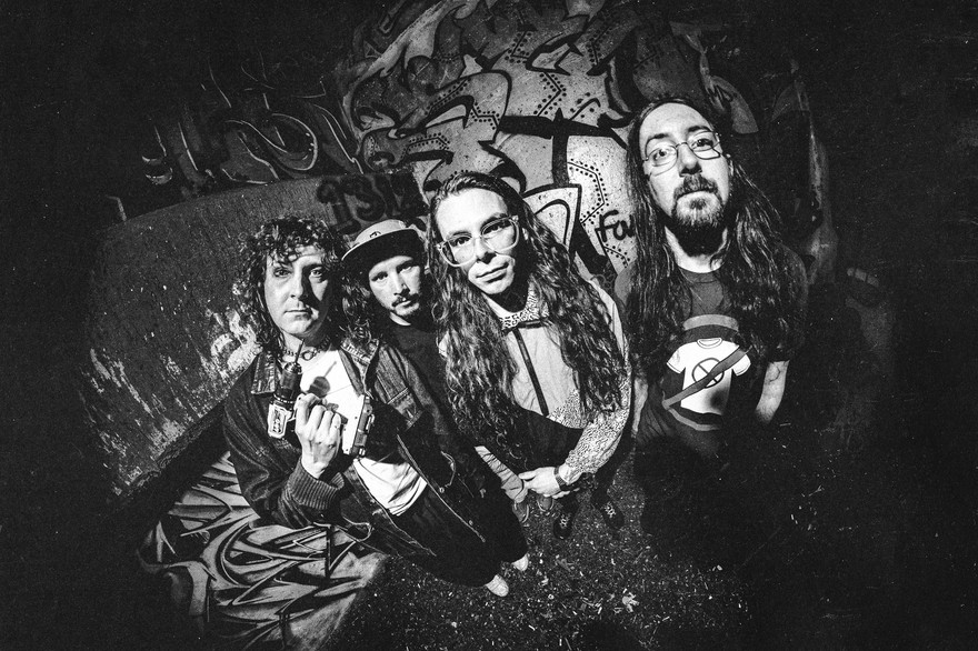 A black and white photo shows the group Tall Mary standing in front of a graffitti wall, each staring into the camera which has a warped vignette effect.