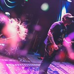 A blurry colour photo shows the group Syphon performing, with two guitar players up front, and the drummer hidden away in the back. The photo is bathed in blue and purple light..