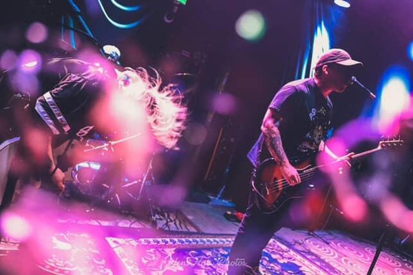 A blurry colour photo shows the group Syphon performing, with two guitar players up front, and the drummer hidden away in the back. The photo is bathed in blue and purple light..