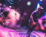 A blurry colour photo shows the group Syphon performing, with two guitar players up front, and the drummer hidden away in the back. The photo is bathed in blue and purple light..