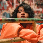 A close-up colour photo shows the artist Tanajah wearing a bright orange coat resting their arms on a metal gate. An out of focus grafitti wall is seen behind them. 