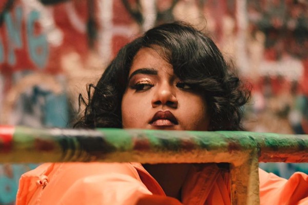 A close-up colour photo shows the artist Tanajah wearing a bright orange coat resting their arms on a metal gate. An out of focus grafitti wall is seen behind them. 