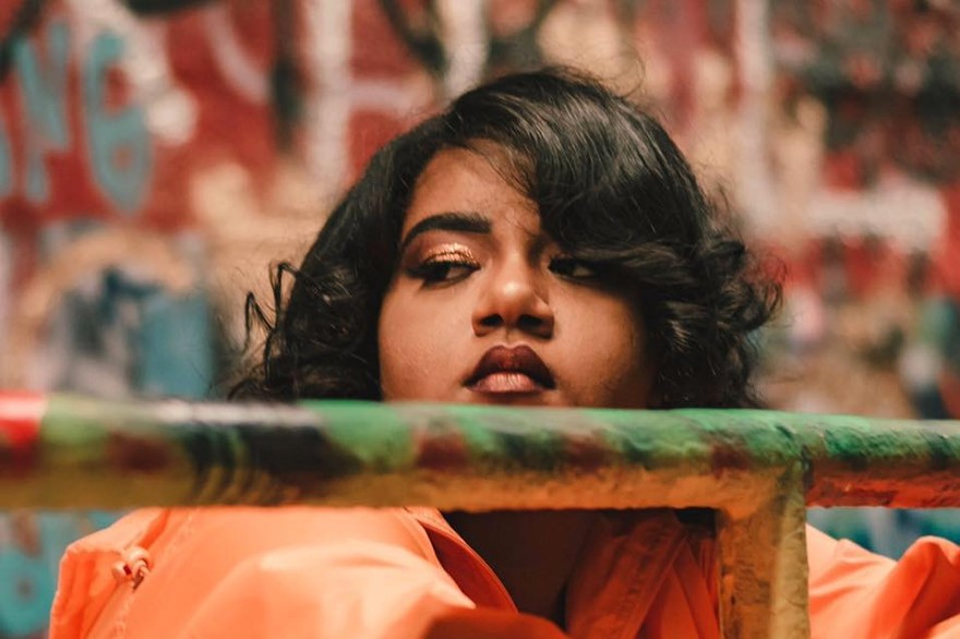 A close-up colour photo shows the artist Tanajah wearing a bright orange coat resting their arms on a metal gate. An out of focus grafitti wall is seen behind them. 