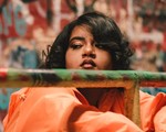 A close-up colour photo shows the artist Tanajah wearing a bright orange coat resting their arms on a metal gate. An out of focus grafitti wall is seen behind them. 