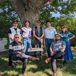 A colour photo taken outdoors shows the seven members of the band Rifle County Players standing on the grass.