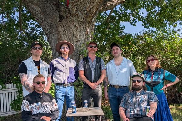 A colour photo taken outdoors shows the seven members of the band Rifle County Players standing on the grass.