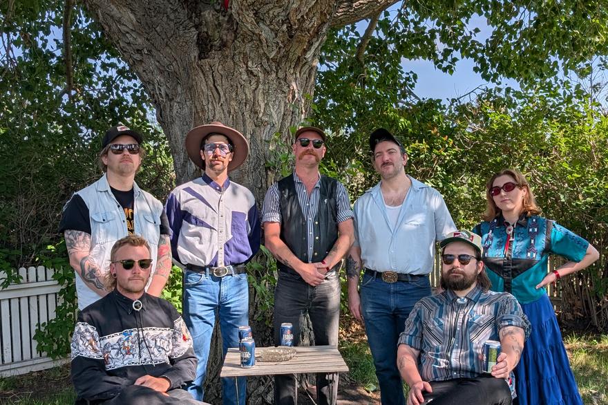 A colour photo taken outdoors shows the seven members of the band Rifle County Players standing on the grass.