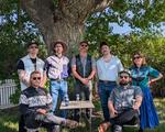 A colour photo taken outdoors shows the seven members of the band Rifle County Players standing on the grass.