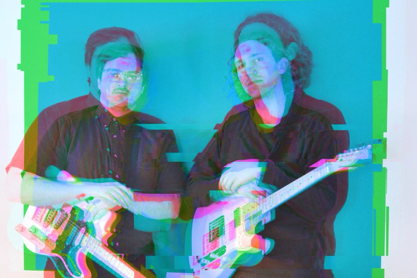 A colour photo with a distorted red and blue 3D type effect shows two members of the band Temps standing side by side wearing all black and both holding white guitars.