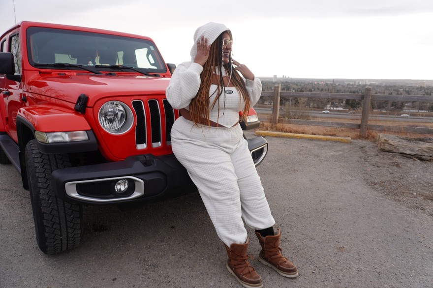 A colour photo shows rapper Tea Fannie standing outside in an all-white outfit leaning against a bright red jeep. 