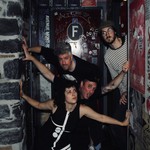 A colour photo of four-piece band Thee Soreheads posing in a doorway with dark brick, red doors and graffiti. 
