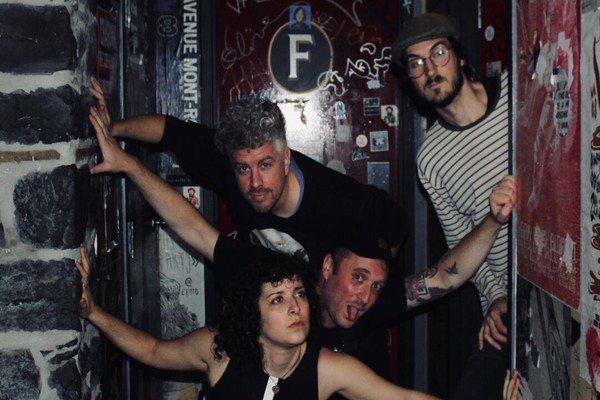 A colour photo of four-piece band Thee Soreheads posing in a doorway with dark brick, red doors and graffiti. 