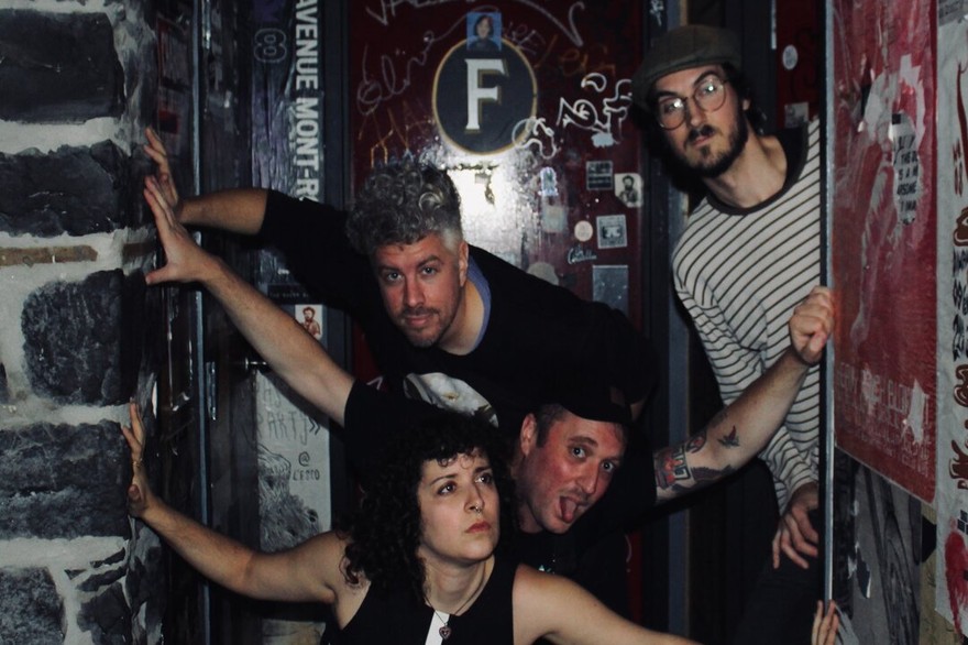 A colour photo of four-piece band Thee Soreheads posing in a doorway with dark brick, red doors and graffiti. 