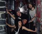 A colour photo of four-piece band Thee Soreheads posing in a doorway with dark brick, red doors and graffiti. 