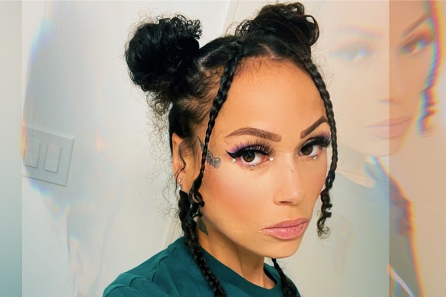 A double exposure photo shows a artist Tinywiings with a close-up on their face, with a faded copy of their face above on the the top right. Their hair is in two tight buns on the top of their head, with two braids hanging down each side. 