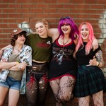 A colour photo shows four-piece Toxic Fem smiling for the camera and standing against a dark brick wall with white spray paint graffiti across it. 