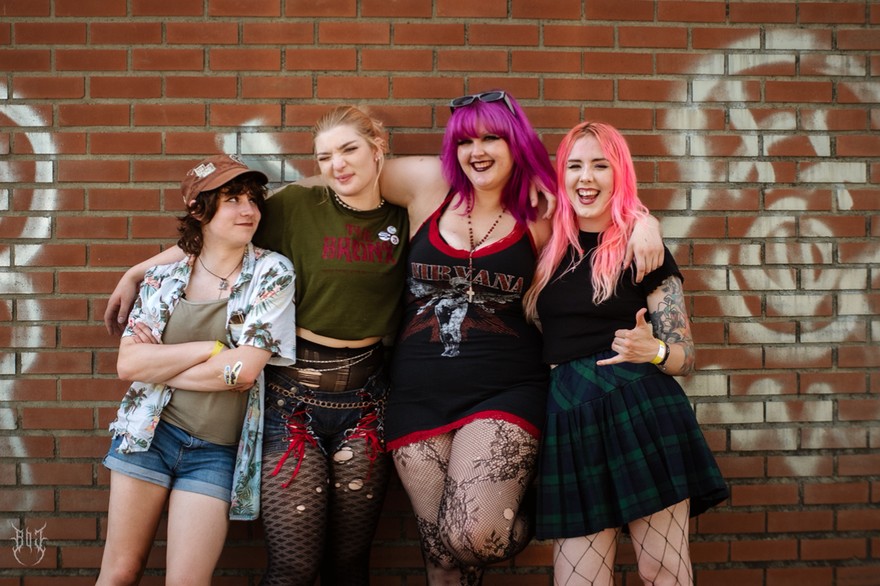 A colour photo shows four-piece Toxic Fem smiling for the camera and standing against a dark brick wall with white spray paint graffiti across it. 