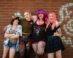 A colour photo shows four-piece Toxic Fem smiling for the camera and standing against a dark brick wall with white spray paint graffiti across it. 