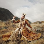 A colour photo shows artist THEIA outside in a field with a renaissance-like orange dress. Their arms are outstretched with pointed fingers. 