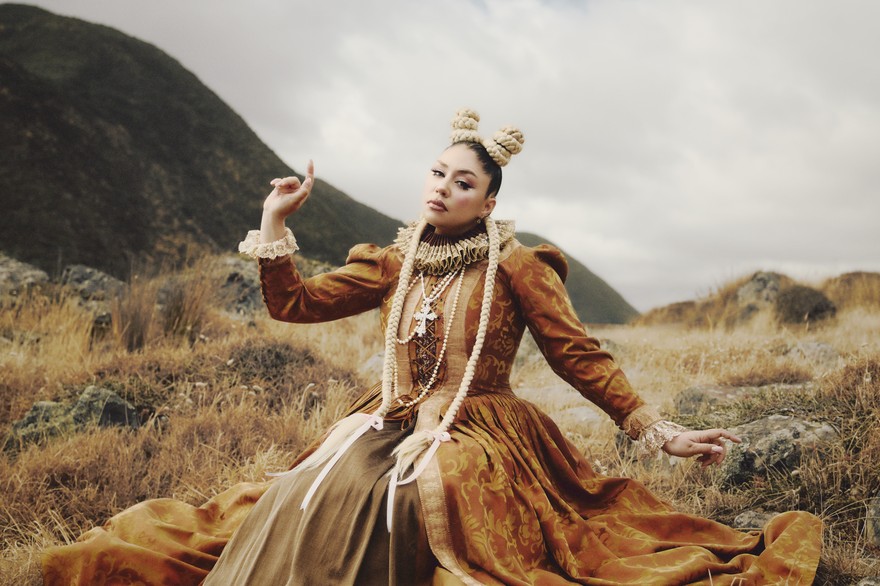 A colour photo shows artist THEIA outside in a field with a renaissance-like orange dress. Their arms are outstretched with pointed fingers. 