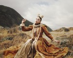 A colour photo shows artist THEIA outside in a field with a renaissance-like orange dress. Their arms are outstretched with pointed fingers. 