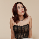 A colour photo shows artist Tricky FM seated on their knees against a beige background. They have a white lacy skirt over fishnet stockings, black heels, a block corset top, and dark brown hair. They;re seen looking up to the top right corner.