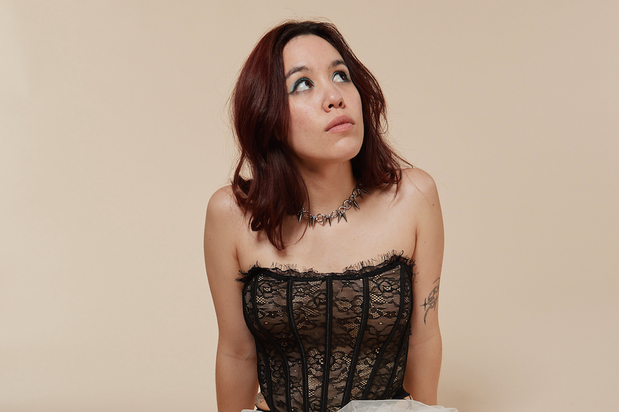 A colour photo shows artist Tricky FM seated on their knees against a beige background. They have a white lacy skirt over fishnet stockings, black heels, a block corset top, and dark brown hair. They;re seen looking up to the top right corner.