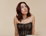 A colour photo shows artist Tricky FM seated on their knees against a beige background. They have a white lacy skirt over fishnet stockings, black heels, a block corset top, and dark brown hair. They;re seen looking up to the top right corner.