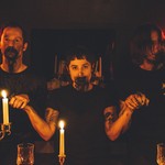 A colour photo shows three-piece group Truster in a seance-like setting. They are bathed in red light, all holding hands, with blood around their mouths. Candles are light on the wood table in front of them, and dim lights are seen in the back. They're all wearing black shirts. 