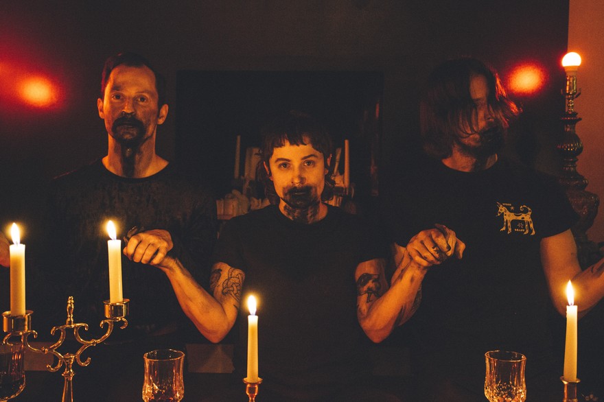 A colour photo shows three-piece group Truster in a seance-like setting. They are bathed in red light, all holding hands, with blood around their mouths. Candles are light on the wood table in front of them, and dim lights are seen in the back. They're all wearing black shirts. 