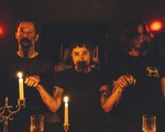 A colour photo shows three-piece group Truster in a seance-like setting. They are bathed in red light, all holding hands, with blood around their mouths. Candles are light on the wood table in front of them, and dim lights are seen in the back. They're all wearing black shirts. 