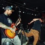 A candid colour photo shows four members of the band VIBES performing on stage. Two guitar players and a bassist are seen, and the singer is flailing mid-kick. The drummer is tucked away in the back. 