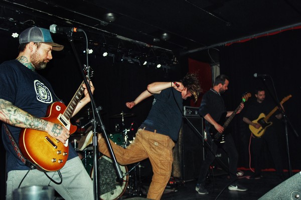 A candid colour photo shows four members of the band VIBES performing on stage. Two guitar players and a bassist are seen, and the singer is flailing mid-kick. The drummer is tucked away in the back. 