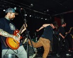 A candid colour photo shows four members of the band VIBES performing on stage. Two guitar players and a bassist are seen, and the singer is flailing mid-kick. The drummer is tucked away in the back. 