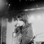 A grainy black and white photo shows the frontperson of the band Uncanny Valley singing into a microphone. They have dark hair, a dark denim jacket and black gloves. 
