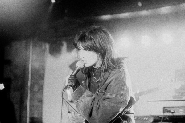 A grainy black and white photo shows the frontperson of the band Uncanny Valley singing into a microphone. They have dark hair, a dark denim jacket and black gloves. 