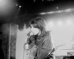 A grainy black and white photo shows the frontperson of the band Uncanny Valley singing into a microphone. They have dark hair, a dark denim jacket and black gloves. 
