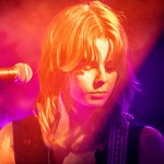 A vibrant colour photo shows a cloe-up of artist Victrix looking down with a microphone in ffront of them. They are bathed in warm purple, red and yellow lights. 