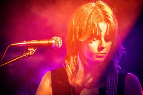 A vibrant colour photo shows a cloe-up of artist Victrix looking down with a microphone in ffront of them. They are bathed in warm purple, red and yellow lights. 