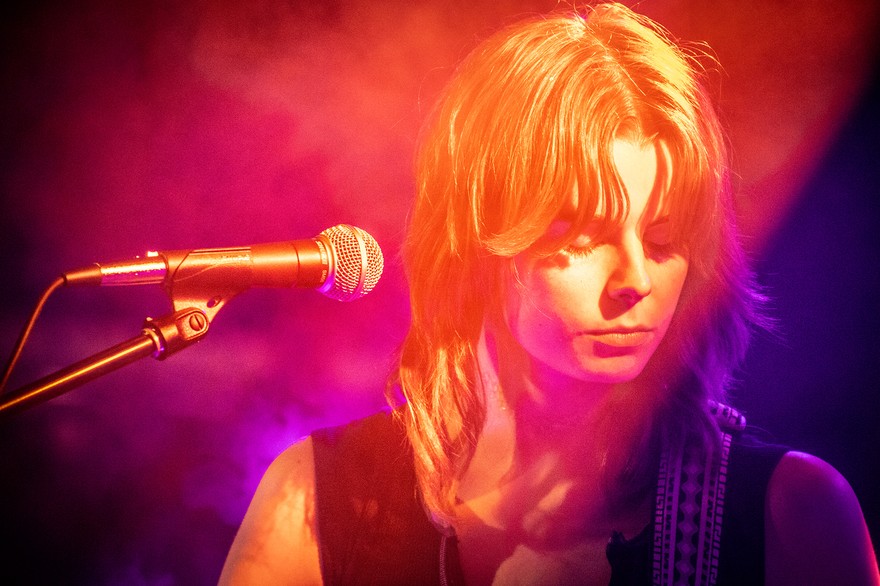 A vibrant colour photo shows a cloe-up of artist Victrix looking down with a microphone in ffront of them. They are bathed in warm purple, red and yellow lights. 