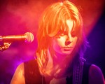 A vibrant colour photo shows a cloe-up of artist Victrix looking down with a microphone in ffront of them. They are bathed in warm purple, red and yellow lights. 