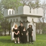 A colour photo shows four-piece band Virgo Rising standing in a grassy green field dressed all in black in front of a white castle-like structure that says "God Loves Us" on the right side. Trees and a body of water are seen in the background. 
