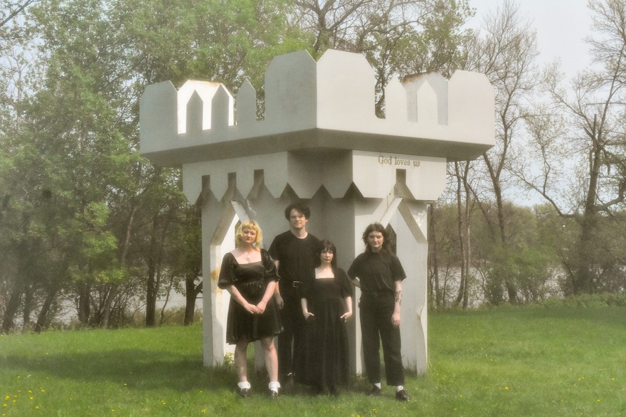A colour photo shows four-piece band Virgo Rising standing in a grassy green field dressed all in black in front of a white castle-like structure that says "God Loves Us" on the right side. Trees and a body of water are seen in the background. 