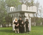 A colour photo shows four-piece band Virgo Rising standing in a grassy green field dressed all in black in front of a white castle-like structure that says "God Loves Us" on the right side. Trees and a body of water are seen in the background. 