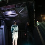 A colour photo shows duo WANTS standing outside at night under a bridge-like structure. Purple light shines from the grate above them. 