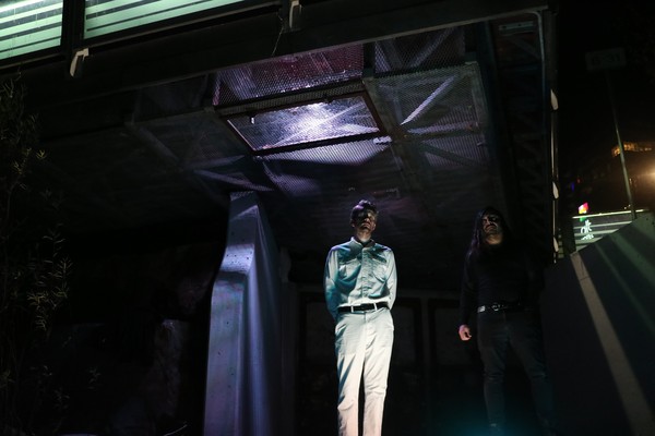 A colour photo shows duo WANTS standing outside at night under a bridge-like structure. Purple light shines from the grate above them. 